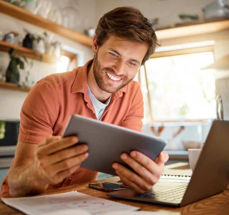 Technology, happy man with tablet and laptop for remote work in kitchen of his home with a lens flare. Social networking or connectivity, online communication and male person smile for email.